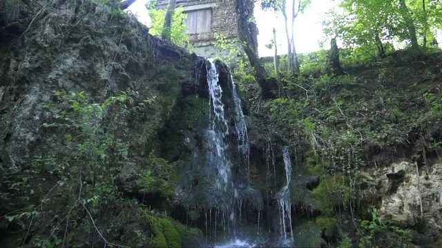 water falls from cliff overgrown with green moss turning into small waterfall against old house and trees on top