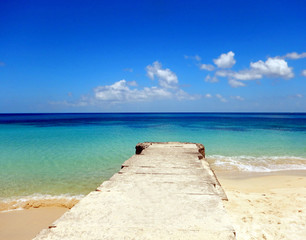 An old, tan stone pier stretching out from a soft, sandy beach into the blue green surf and the empty, calm indigo sea beyond with a bright blue sky scattered in white, puffs of clouds.