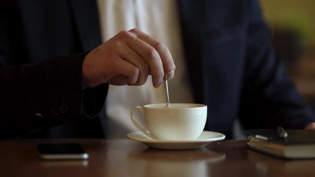 Strong man hands stirring coffee/latte/cappuccino with teaspoon in a cafe. Man dressed in classic black suit and whire shirt. There are pen, notebook, phone on table. Close up faceless concept