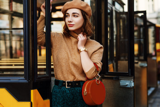Outdoor portrait of young fashionable woman wearing beige beret, turtleneck, green corduroy trousers, wrist watch, holding round orange suede handbag, posing in street of european city. Copy space