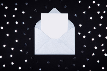 White blank card in opened silver grey envelope on black background decorated with star confetti. 