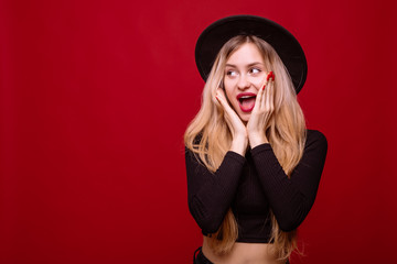 Young blonde woman in fluppy black hat and black clothes surprised isolated on red background