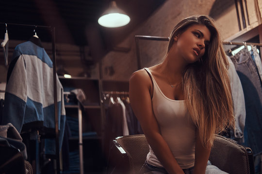 Seductive girl wearing a white t-shirt and jeans sitting on a chair and looking away in the fitting room of a clothing store.