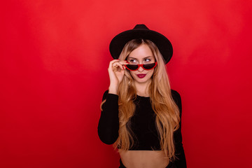 Fashion portrait pretty woman in black style hat and sunglasses over red background. Holiday concept