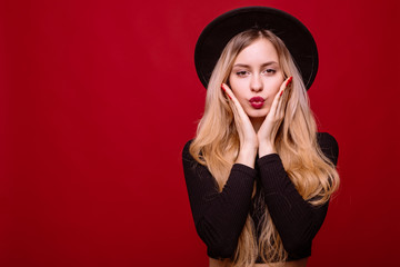 Young blonde woman in fluppy black hat and black clothes kiss on camera isolated on red background