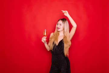 Portrait of an excited young woman dressed in red dress holding glass of champagne and celebrating isolated over red background