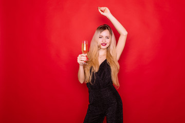 Portrait of a cheerful young blonde woman in dress holding glass of champagne toasting camera isolated over red background.