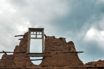 Brick wall with adobe clay plaster and broken wooden window frame remained of ruined old rural country house on cloudy blue sky background