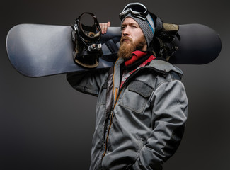 Confident man with a red beard wearing a full equipment holding a snowboard on his shoulder, looking away with a serious look, isolated on a dark background.