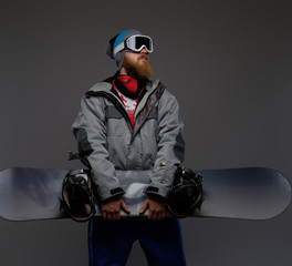 Brutal man with a red beard wearing a full equipment holding a snowboard and looking away, isolated on a dark background.