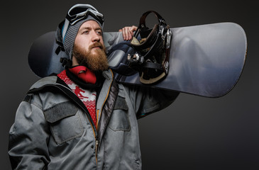 Confident man with red beard wearing a full equipment holding a snowboard on his shoulder, isolated on a dark background.