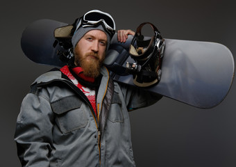 Confident man with red beard wearing a full equipment holding a snowboard on his shoulder, isolated on a dark background.