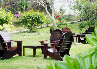A grouping of glossy, varnished wooden Adirondack chairs and tables sitting on a grassy hillside surrounded by lush, tropical plants with a path and stone wall in the background.