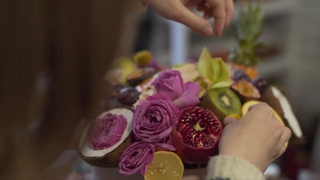 Close-up scene oa a wonderful bouquet from violet and orange roses, different fruits and coconut. The bunch of flowers is being made by florist in flower shop.