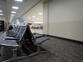 Row of empty sleek silver metal and black leather arm chairs lined up for waiting passengers at an airport gate, with grey and beige abstract carpeting, travelers and monitor screens in background.