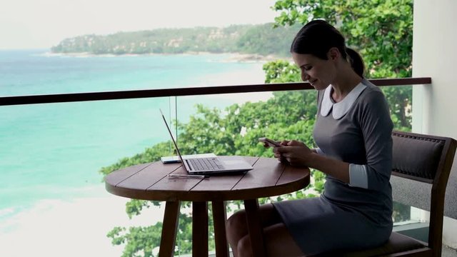 Young businesswoman sitting with smartphone on terrace, with sea view, 4K

