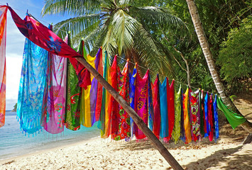 Colorful fabric scarves or cloth sarongs printed with beautiful tropical designs hanging in a line and held up by a bamboo pole over a sunny palm tree lined beach in front of calm aquamarine water.