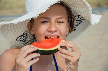 A young girl in a white straw hat is eating red, ripe watermelon on the beach on a hot day with an...