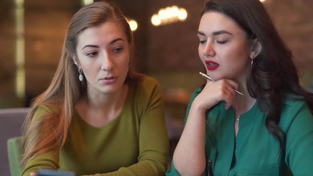 Two attractive young women are sitting in a cafe indoors and talking in a video chat with somebody by phone. One girl is holding a phone and another is writing something down in her notebook.