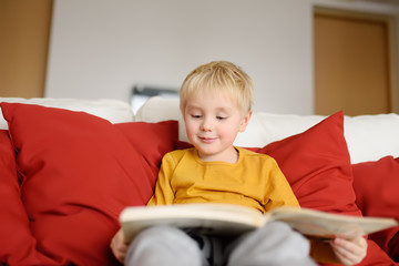 Fototapeta premium Little boy is sitting at home on the couch and reading a book. Learning to read.