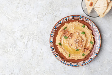 Hummus in to a brown clay plate with a blue pattern. On the white table are vegetables, greens, triangular pieces of pita. Top view. Flat lay.