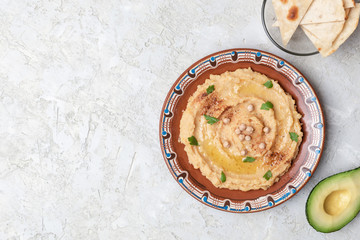 Hummus in to a brown clay plate with a blue pattern. On the white table are vegetables, greens, triangular pieces of pita. Top view. Flat lay.