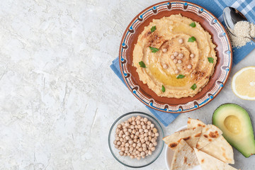 Hummus in to a brown clay plate with a blue pattern. On the white table are vegetables, greens, triangular pieces of pita. Top view. Flat lay.