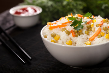 Cooked white rice with vegetables in a bowl with sauces and chopsticks on the wood black bamboo background.