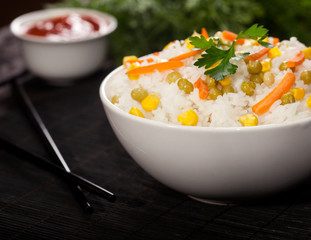 Cooked white rice with vegetables in a bowl with sauces and chopsticks on the wood black bamboo background.