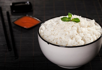 Cooked white rice in a bowl with sauces and chopsticks on the wood black bamboo background.