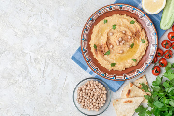 Hummus in to a brown clay plate with a blue pattern. On the white table are vegetables, greens, triangular pieces of pita. Top view. Flat lay.