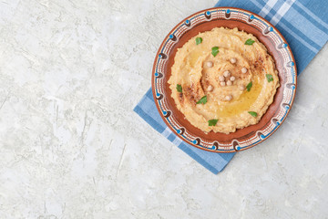 Hummus in to a brown clay plate with a blue pattern. On the white table are vegetables, greens, triangular pieces of pita. Top view. Flat lay.