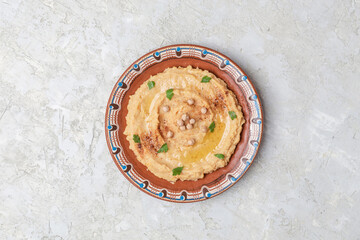 Hummus in to a brown clay plate with a blue pattern. On the white table are vegetables, greens, triangular pieces of pita. Top view. Flat lay.