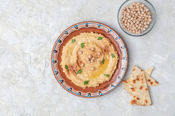 Hummus in to a brown clay plate with a blue pattern. On the white table are vegetables, greens, triangular pieces of pita. Top view. Flat lay.