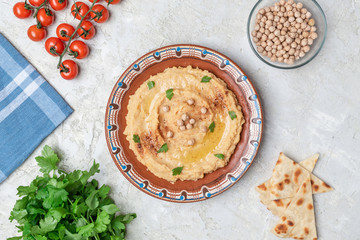 Hummus in to a brown clay plate with a blue pattern. On the white table are vegetables, greens, triangular pieces of pita. Top view. Flat lay.