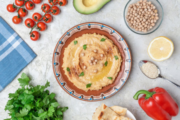 Hummus in to a brown clay plate with a blue pattern. On the white table are vegetables, greens, triangular pieces of pita. Top view. Flat lay.