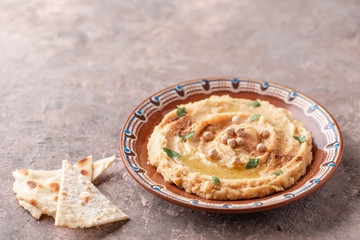 Hummus in a brown clay plate with a blue pattern. On the brown table are vegetables, greens, triangular pieces of pita. 