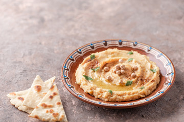 Hummus in a brown clay plate with a blue pattern. On the brown table are vegetables, greens, triangular pieces of pita. 