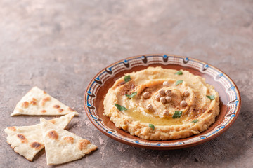 Hummus in a brown clay plate with a blue pattern. On the brown table are vegetables, greens, triangular pieces of pita. 