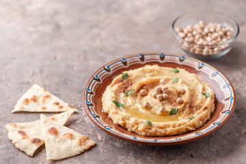 Hummus in a brown clay plate with a blue pattern. On the brown table are vegetables, greens, triangular pieces of pita. 