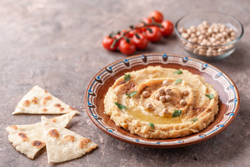 Hummus in a brown clay plate with a blue pattern. On the brown table are vegetables, greens, triangular pieces of pita. 