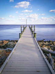 Fototapeta na wymiar Colorful, Serene Photo of a Dock Extending Out To the Ocean - with a Patch of Rocks and Logs, and Calm Waters on a Bright, Mildly Cloudy Day in the Mid Atlantic United States