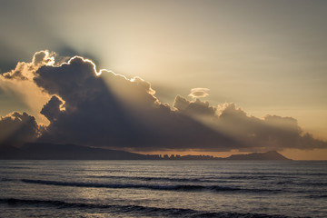 Naklejka na ściany i meble Ocean on a Cloudy Sunrise - with Rays of Sun Shining Around the Edge of the Clouds on the Horizon, and Calm, Empty Waves Below on a Summer Morning in Hawaii