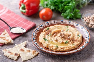 Hummus in a brown clay plate with a blue pattern. On the brown table are vegetables, greens, triangular pieces of pita. 