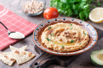 Hummus in a brown clay plate with a blue pattern. On the brown table are vegetables, greens, triangular pieces of pita. 