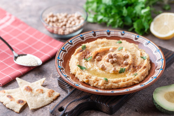 Hummus in a brown clay plate with a blue pattern. On the brown table are vegetables, greens, triangular pieces of pita. 