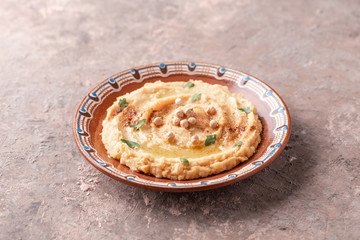 Hummus in a brown clay plate with a blue pattern. On the brown table are vegetables, greens, triangular pieces of pita. 