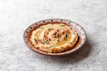 Hummus in to a brown clay plate with a blue pattern. On the white table are vegetables, greens, triangular pieces of pita. Top view. Flat lay.
