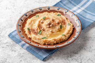 Hummus in to a brown clay plate with a blue pattern. On the white table are vegetables, greens, triangular pieces of pita. Top view. Flat lay.