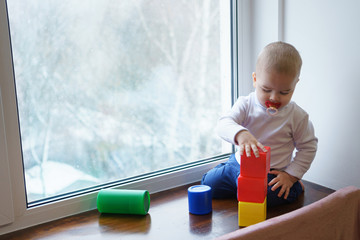 Little boy with dummy sits on windowsill. He builds tower of cubes. Winter day outside window.